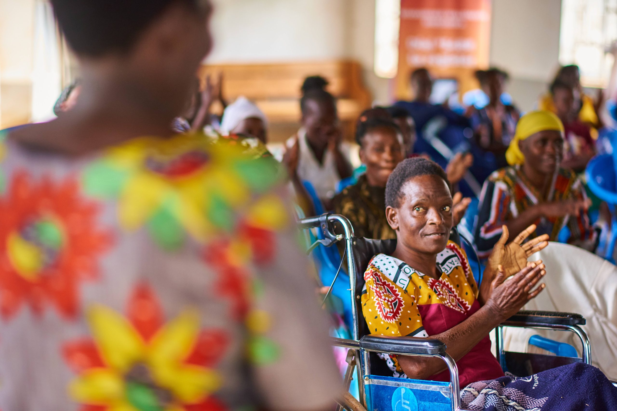 A woman using a wheelchair claps while seated among a group of women in a community meeting, facing a facilitator in the foreground.